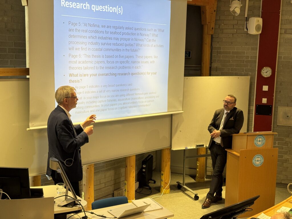 Audun Iversen i samtale med førsteopponent Jesper Raakjær under sin disputas. De to står foran i en amfisal.