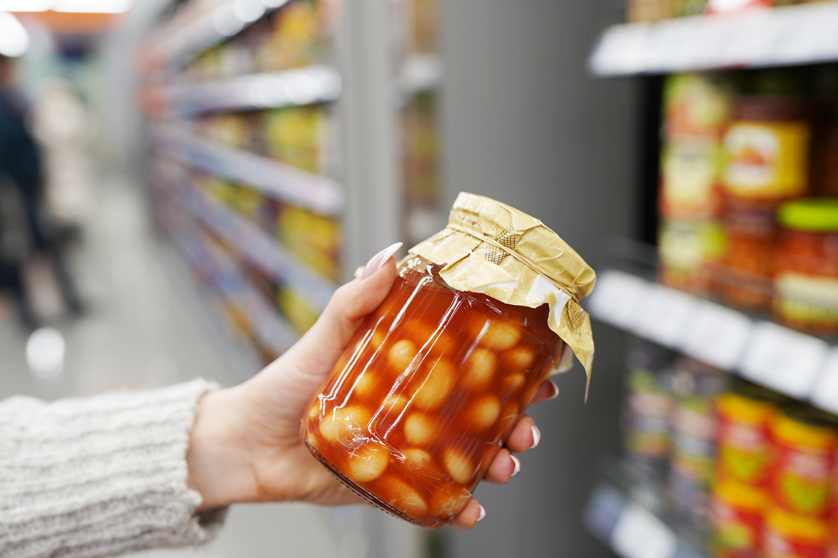Bildet viser en person som holder glass med hermetiserte bønner i tomatsaus i butikk.